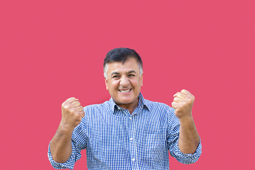 Portrait of cheering mature man agaist colored background. Man celebrating his success and looking away with positive expression. He wearing shirt. Standing in front of colored background. Studio shot and developed from Raw format. Horizontal composition.