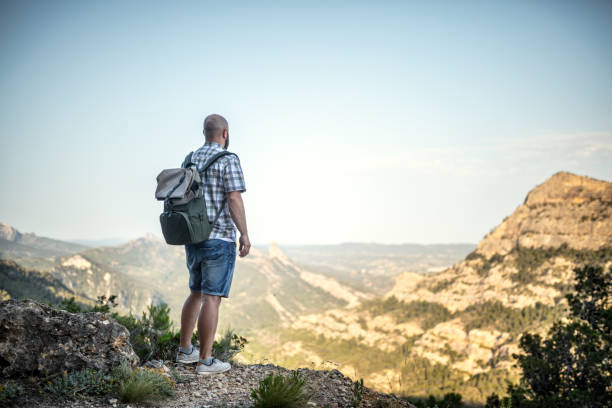 Traveler with backpack stands on a huge rock and looks on a mountain peak in front of him. Overcast sky.