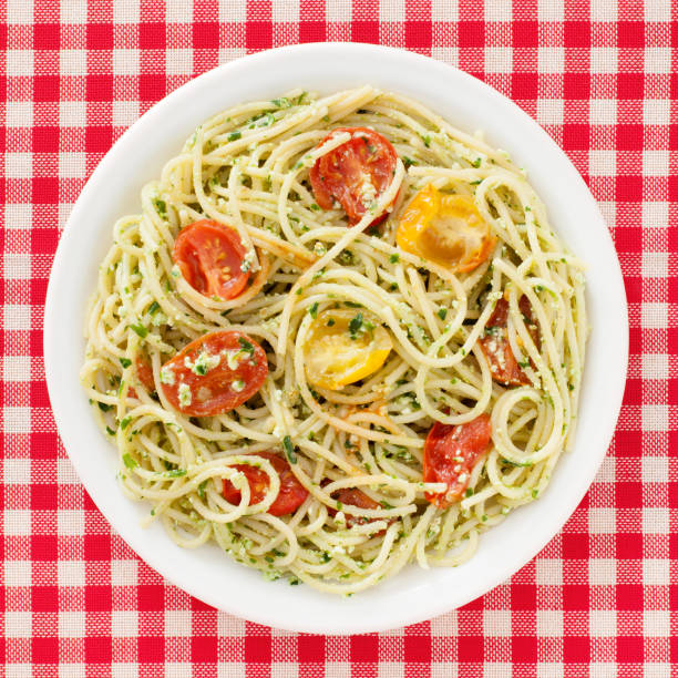 Top view of white dish over checked red surface with pasta consisting of spaghetti with ricotta, parsley pesto and cherry tomatoes