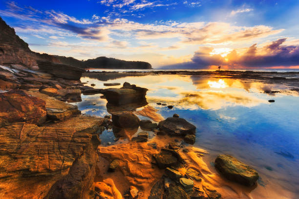 Reflection of clouds and rising sun in still salt water puddle during low tide sea bed opening near sandstone cliffs of Narrabeen headland in Sydney's pacific coast.