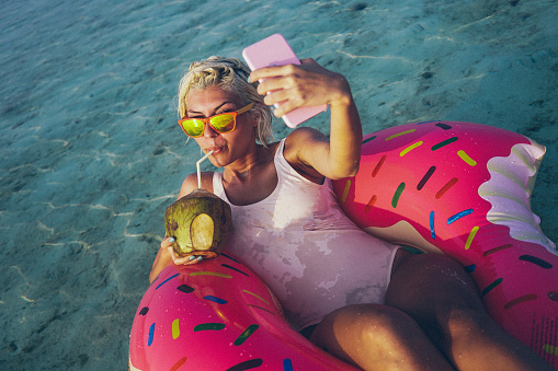 Young, attractive and beautiful woman is taking a selfie while floating in float in the ocean. Float is pink and yellow and have shape of doughnut witch is missing one part, something like one bite. She is wearing white swimsuit and orange sunglasses. In her left hand she is holding her smart phone while coconut fruit is in right hand and she is drinking tasty coconut juice from this big fruit. Smile on her face is sign that she is relaxing and enjoying in this quite environment. Her short blonde hair is wet. Water is crystal clear and turquoise colored.