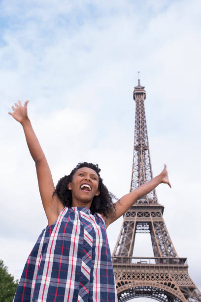 Excited woman tourist visiting Eiffel Tower. Woman laughing, standing near to tower with arms raised and wide open. Champ de Mars, Paris, France.