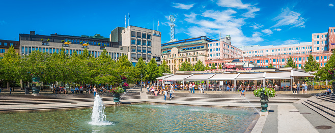 Crowds of shoppers, locals and tourists enjoying the summer sunshine beside the fountains of Kungstradgarden overlooked by the department stores of Hamngatan shopping street in the heart of Stockholm, Sweden's vibrant capital city.