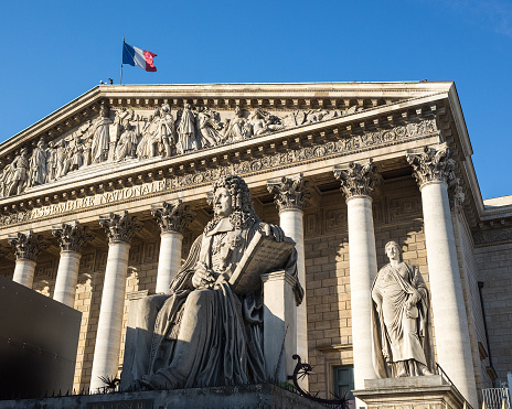Facade with colonnade of the Palais Bourbon in Paris, seat of the french National Assembly, with the statue of Francois d'Aguesseau and the french flag flying on the roof against blue sky.