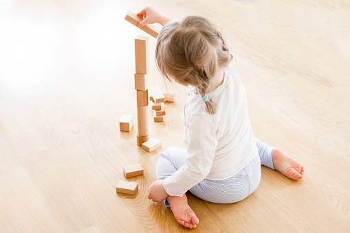 Little girl (two years old) is sitting on the floor and playing with her building blocks. She is building a high tower and are just about to put another block to it. She is sitting with her back against the viewer, totally lost in her play.