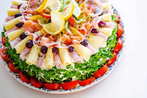 A fresh, colorful and homemade savory sandwich cake (called ”Smörgåstårta” in Sweden) on white table. It is a cake where the cake layers are made of sandwiches that has savory fillings and mixed cheese, ham, fruits and vegetables on top. Traditionally eaten in summertime celebrations such as birthdays or baptisms.
