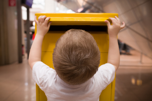 A little blond boy in a white t-shirt puts a letter into a yellow postbox. Sending post to a friend at the post office.