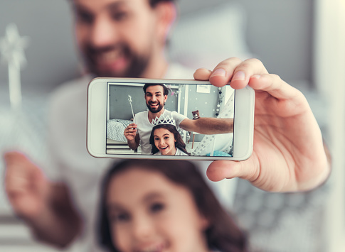 Cute little daughter and her handsome young dad in crowns are doing selfie using a smart phone and smiling while playing together in child's room