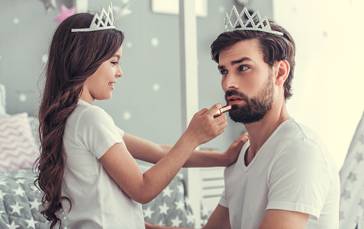 Cute little daughter and her handsome young dad in crowns are playing together in child's room. Girl is doing her dad a makeup