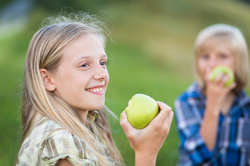 Young girl portrait eating apple. Her sibling in the background. both blond, both smiling.