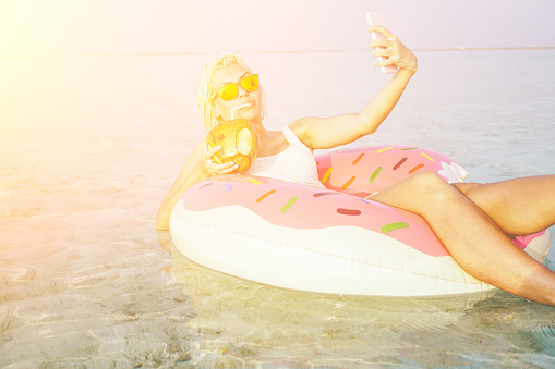 Young, attractive and beautiful woman is taking a selfie while floating in float in the ocean. Float is pink and yellow and have shape of doughnut witch is missing one part, something like one bite. She is wearing white swimsuit and orange sunglasses. In her left hand she is holding her smart phone while coconut fruit is in right hand and she is drinking tasty coconut juice from this big fruit. Smile on her face is sign that she is relaxing and enjoying in this quite environment. Her short blonde hair is wet. Water is crystal clear and turquoise colored.