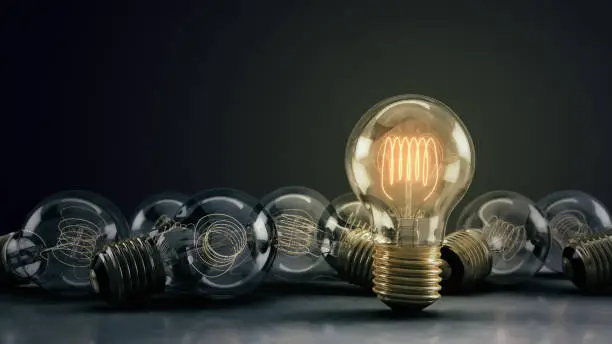 Multiple incandescent light bulbs on a reflective surface and dark background.  One hero bulb illuminated, standing out from the rest.  Great for conveying a big idea or unique business thoughts.  3D illustration.