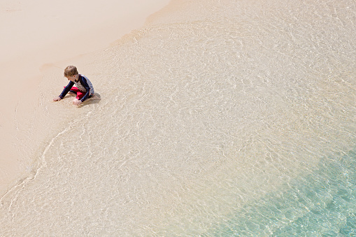 aerial view of little boy in swimwear playing with sand at the beach by water, copyspace on right, vacation concept