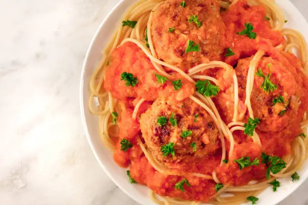 A closeup of a plate of meatballs with pasta, tomato sauce, and fresh parsley, shot from above on a white marble texture with a place for text