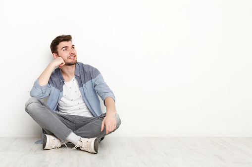 Pensive young man sitting on floor and looking upwards. Handsome relaxed guy dreaming about something, lost in thoughts, isolated on white background