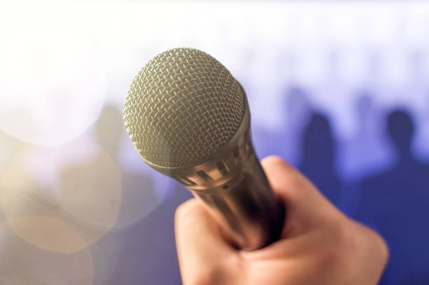 istock Public speaking and giving speech concept. Hand holding microphone in front of a crowd of silhouette people with lens flare and sun light leak bokeh. Singing to mic in karaoke or talent show concept. 813024546
