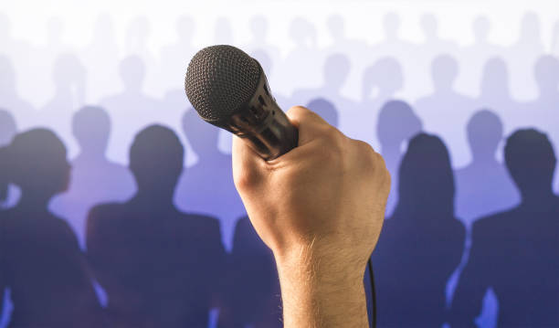 istock Public speaking and giving speech concept. Hand holding microphone in front of a silhouette audience and crowd of people. Singing to mic in karaoke or talent show concept. 813024552