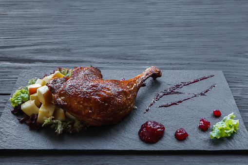 Roasted duck leg closeup served with apples and cherry sauce. Restaurant food on black slate plate at wood table