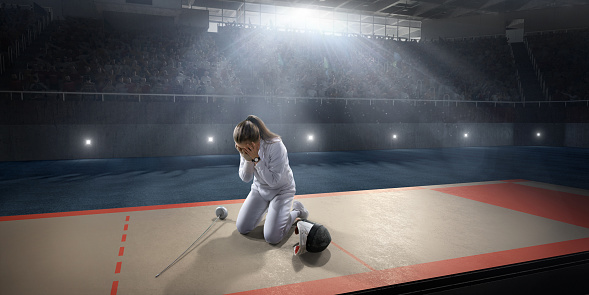 Sad fencer stands on the big professional stage. The fencer lost the competition. She is wearing an unbranded fencing suit. The fencer holds a sword and protective helmet.