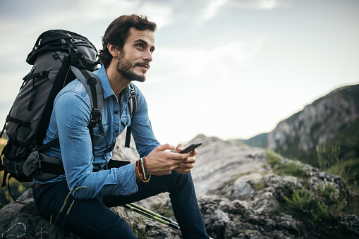 Young handsome hiker using mobile phone while resting on the top of a mountain