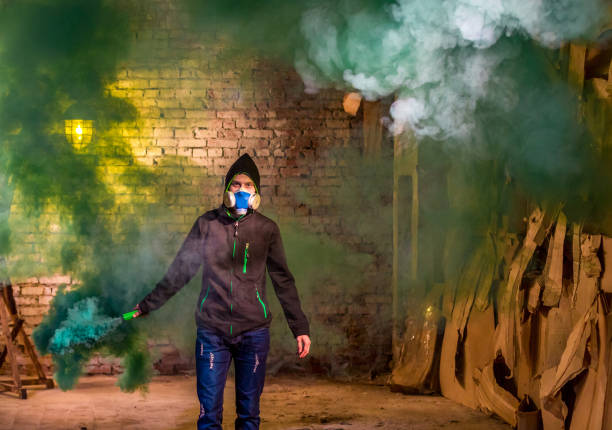 The protester young man dressed in the dark hoodie in protective gas mask on his face. The young guy is holding the color smoke bomb in his hand. He is looking at the camera. Shooting in abandoned building at a urban slums