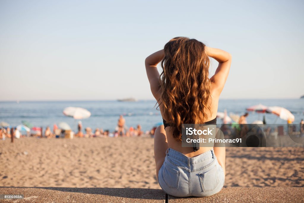 Sun is up, move your body! Beautiful brunette girl holding her hair up and sitting on the sandy beach Hair Stock Photo