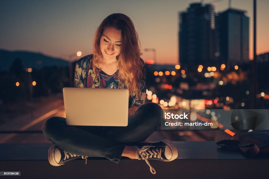 Teenage girl using laptop by night against the urban skyline Young girl sitting at the bridge and surfing the net on laptop Laptop Stock Photo