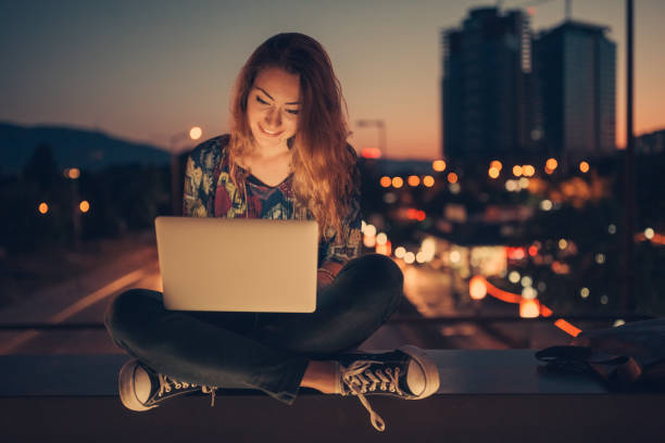 Teenage girl using laptop by night against the urban skyline Young girl sitting at the bridge and surfing the net on laptop teenage girls dusk city urban scene stock pictures, royalty-free photos & images