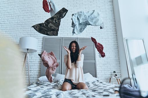 Beautiful young woman sitting on bed and making face while clothing flying everywhere