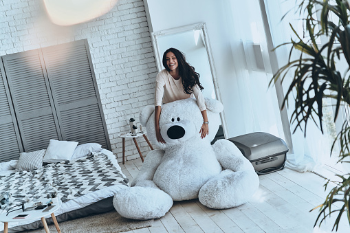 Beautiful young woman touching large teddy bear and smiling while standing in the bedroom at home