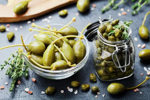 Mixed capers in jar and bowl on black stone kitchen table.