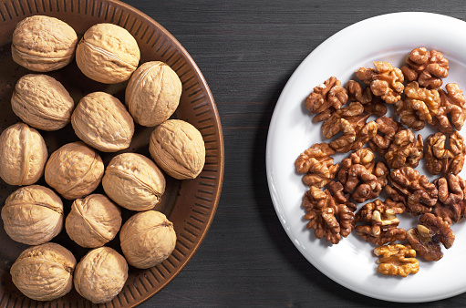 Walnuts whole and kernels in plate on dark wooden table, top view