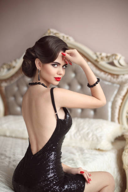 Beautiful stunning elegant lady in black glitter sequins dress sitting on luxury sofa in modern interior. Fashion gems jewelry set. Bright red lips hollywood make up.