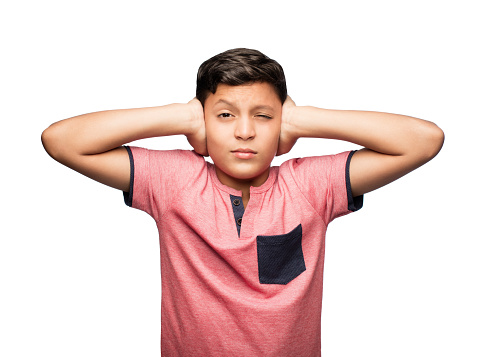 A latin teenage boy covering his ears, looking at the camera and squinting.