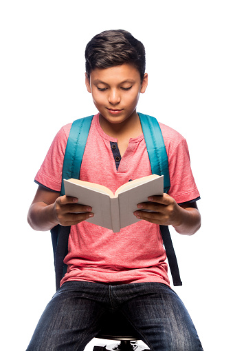 A latin teenage schoolboy stitting, holding a book and reading it.