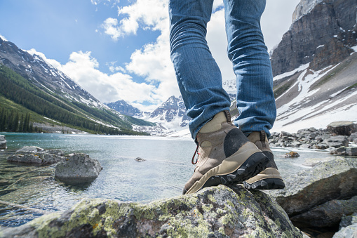 Low angle view of hiker standing on rock above mountain lake, close up on hiking boots, sunbeam.