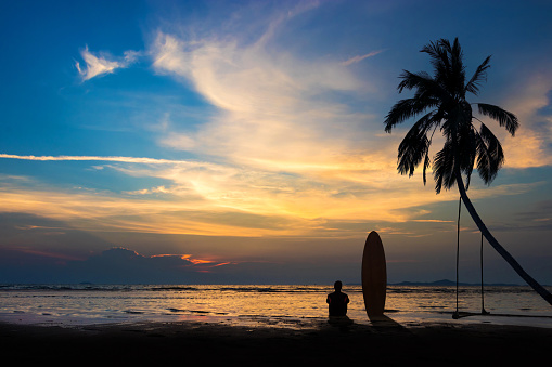 Silhouette of surf man sit with a surfboard on the beach. Surfing scene at sunset beach with colorful sky. Outdoor water sport adventure lifestyle.Summer activity. Handsome Asia male model in his 20s.