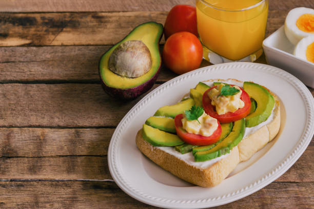 istock Open sandwich style for breakfast or lunch. Sandwich spread with cream cheese, avocado, tomato and boiled eggs on white plate. Avocado open sandwich style serve with orange juice on rustic wood table. 813533400