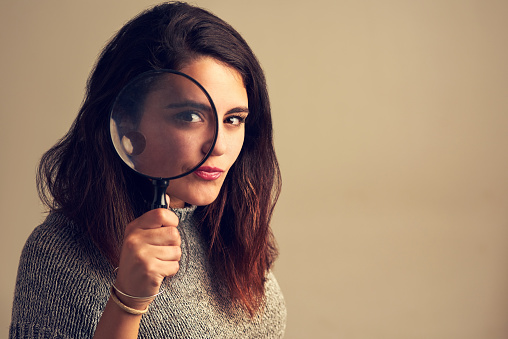 Studio portrait of a young woman looking through a magnifying glass against a brown background