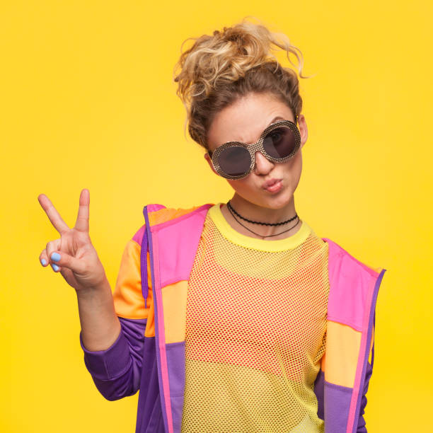 Hipster girl in colorful jacket making v-sign Blonde hipster girl with hair bun wearing colorful jacket, yellow sheer shirt grimacing making v-sign. cool attitude fashion stock pictures, royalty-free photos & images