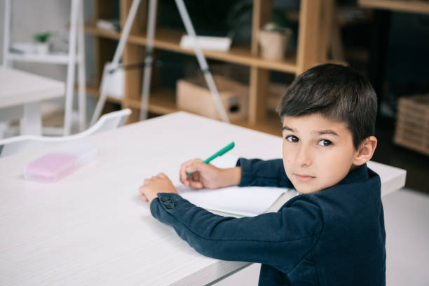istock Cute serious schoolboy writing in exercise book while sitting at desk and looking at camera 813769238