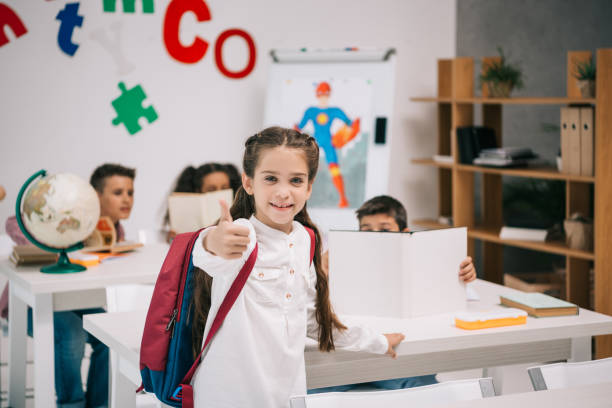 istock Cute smiling schoolgirl showing thumb up while classmates studying behind 813779778