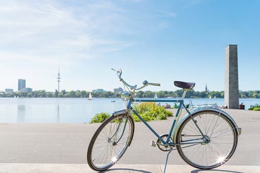 vintage blue bicycle parked at lakeshore of Alster Lake in Hamburg, Germany under beautiful clear blue summer sky with cityscape in blurred background