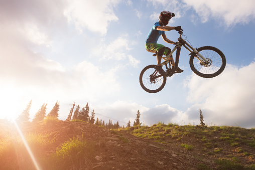 Low angle view of man catching air with dirt bike while riding downhill rough terrain from mountain plateau illuminated by bright sunray shinning from behind during the golden hour
