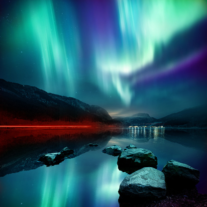 A large Northern Lights (aurora borealis) display glowing over a mountain pass and reflected on a lake at night. Photo composition.