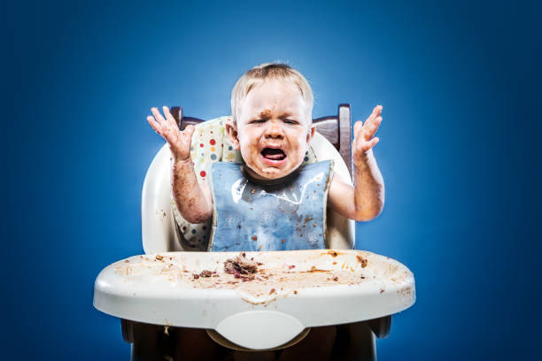 A one year old child sitting in a highchair and covered with his meal.  A typical day in life of a baby.