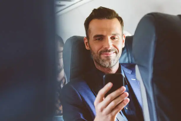 Close up of happy mature man travelling by plane and using mobile phone. Male passenger with smart phone during flight.