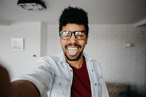 African ethnicity man at the apartment is using smartphone to take photos of himself. Smiling mixed race person using phone to take selfies inside the living room. Man is making various facial expressions while doing selfie.