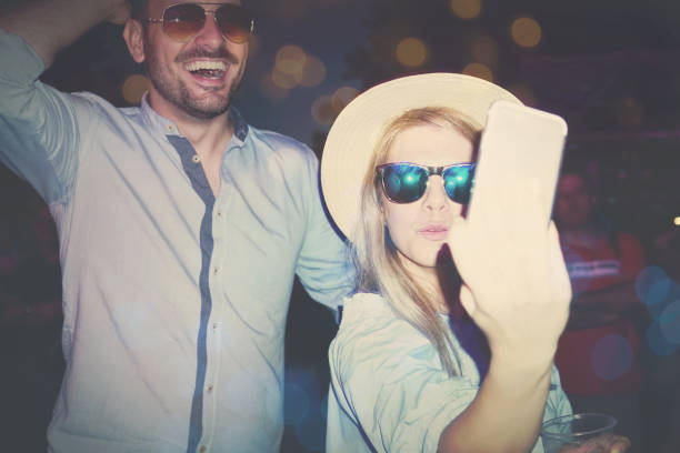 Young and attractive couple are taking selfie on festival. They are having great time together with their favorite performers and they need to catch that moment for memory. Both are dressed casual so they are ready to dance and jump all night till morning. Both have sunglasses and girl have hat on head.