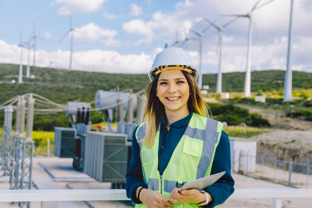 Portrait of young confident female engineer in electric power station Portrait of young happy electrical engineer and technician worker young woman look at camera posing while standing, smiling, working with digital tablet pc in front of electrical components, wind turbines and towers of high tension of an electricity power generation station field in a sunny day rural landscape. xxxl size power plant workers stock pictures, royalty-free photos & images